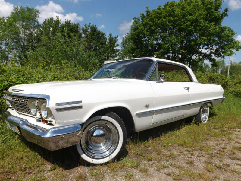 Dieses Foto zeigt den Chevrolet Impala, einen Oldtimer aus dem Jahr 1963, der aktuell bei My Next Classic zum Verkauf angeboten wird. Das Sportcoupé mit V8-Motor, Servolenkung und -bremse ist in einem sehr guten Zustand und hat eine tolle Farbkombination aus weiß und Innenausstattung in Vollleder rot.